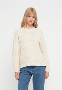 Chunky knit cream sweater with ribbed texture, crew neckline, and dropped shoulders. Paired with light blue denim jeans.