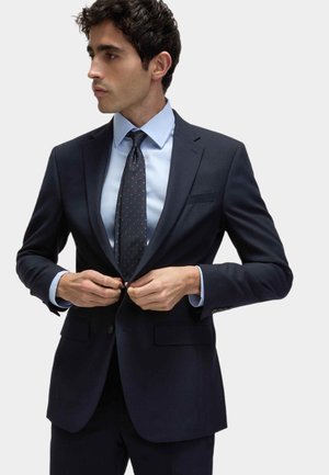Dunkelblauer Anzug mit slim fit, ausgestattet mit zwei Knöpfen, Reverskragen und Taschen-details. Hellblaues Hemd und dunkel gemusterte Krawatte vervollständigen den Look.
