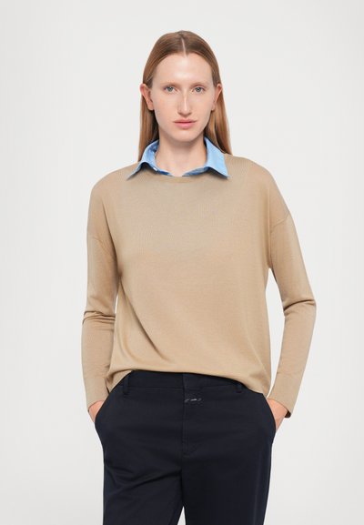 Beige knitted sweater with a round neckline worn over a light blue collared shirt, paired with dark navy trousers. Smooth texture, relaxed fit.