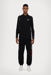 Polo shirt nero a maniche lunghe con logo, abbinato a pantaloni sportivi neri con pannelli grigi e una patch grafica arancione, indossato con sneakers bianche.