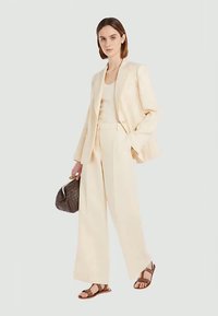 Cream-colored ensemble featuring a tailored blazer and wide-leg trousers, paired with a fitted tank top and brown sandals, carrying a textured handbag.