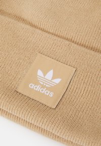 Hnedý pletený čiapka s textúrovaným vzorom, so všitou štvorcovou nášivkou zobrazujúcou biele logo Adidas a tri pruhy.