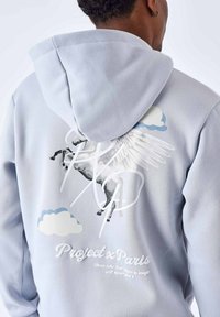 Sweat à capuche gris avec un grand graphique d'un cheval en train de sauter avec des ailes, accompagné de nuages et de texte au dos. Matière en coton, coupe décontractée.