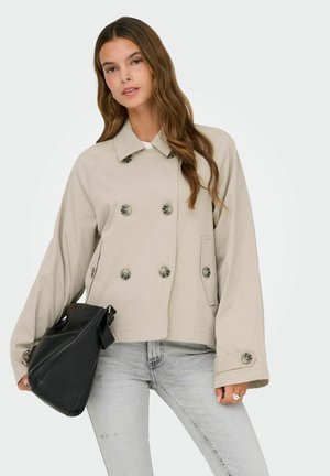 Fiatal nő, bézs színű, dupla soros zakót és világosszürke farmert visel, fekete kézitáskát tart, hosszú, hullámos hajjal.