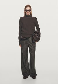 Ciemnobrązowy prążkowany golf z akcentami na ramionach, w połączeniu z luźnymi szarymi spodniami i ciemną skórzaną torbą. Modelka w okularach przeciwsłonecznych.