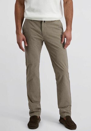 Mann trägt beige Slim-Fit-Hose, weißes Hemd und dunkelbraune Loafers, steht vor einfarbig hellem Hintergrund.
