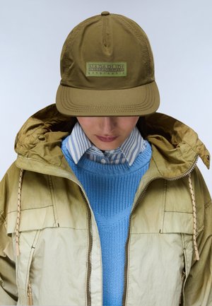 Personne portant une casquette Napapijri vert olive, une veste à capuche vert olive par-dessus un pull en maille bleu et une chemise rayée bleue et blanche avec col.