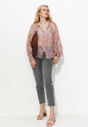 Femme portant un chemisier floral transparent rose, un jean gris, des sandales dorées et tenant un sac à franges marron, se tenant devant un fond blanc.