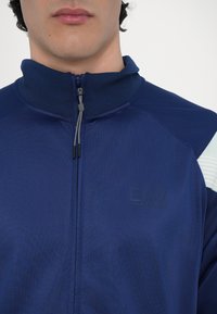 Giacca zip con colletto alto in tessuto testurizzato di colore navy. Presenta un logo sul petto e dettagli a righe blu chiaro sulle spalle.