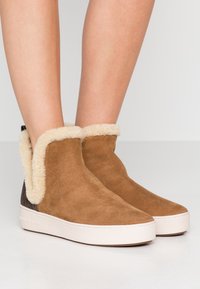 Bottines en suede marron avec un bord en fausse fourrure crème, semelle blanche et côtés texturés, conçues pour le confort et la chaleur.