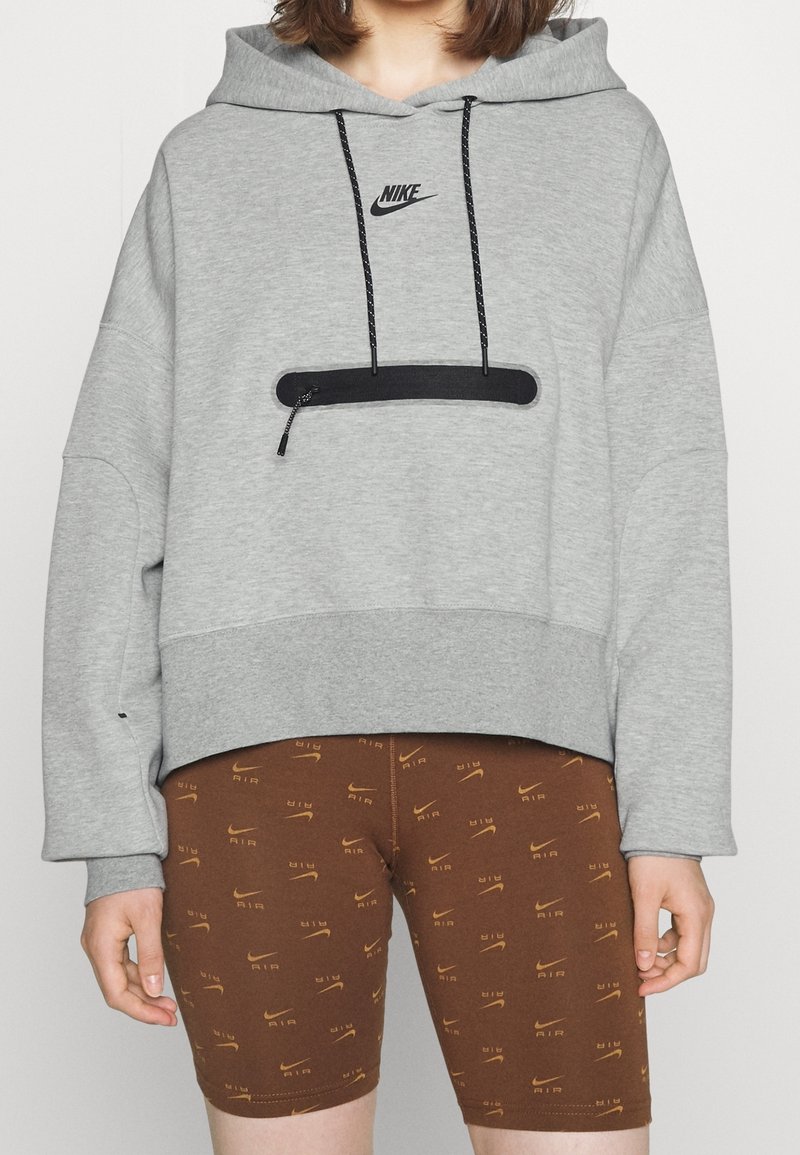 Sudadera gris con un bolsillo frontal y cremallera negra, con un pequeño logo de Nike. Combinada con pantalones cortos marrones con un patrón de logo repetido.