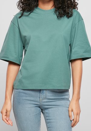 Vrouw die een loszittend, groen T-shirt met korte mouwen en lichtblauwe jeans draagt, staand tegen een effen grijze achtergrond.
