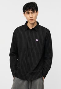 Νεαρός άνδρας που φοράει μαύρο πουκάμισο με κάθετες λεπτές ρίγες και μικρό μωβ ταμπελάκι με λογότυπο στο στήθος, με τα χέρια στις τσέπες του γκρι παντελονιού.