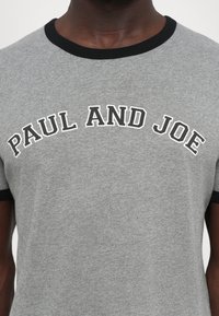 Temna oseba z zdravo kožo, oblečena v sivo majico s črnimi robovi in belim napisom "PAUL AND JOE" čez prsa.