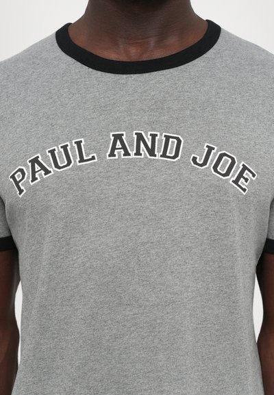 Tume nahk inimene, kellel on seljas hall T-särk mustade ääristega, millel on valge ääristusega tekst "PAUL AND JOE" rinna peal.