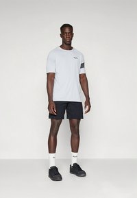 Camiseta de manga corta gris claro con un logo negro, combinada con pantalones cortos azul marino. El modelo lleva zapatillas negras y calcetines blancos. Diseño sencillo.