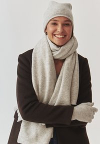 Hallitud kootud müts, sall ja kinda pehmest villast segu; mudel kannab tumedat mantel; sall keerab kaela ümber, näidates peent tekstuuri.