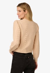 Beżowy sweter dziergany z bufiastymi długimi rękawami, ozdobiony wzorem liści na plecach, zestawiony z czarnymi spodniami z wysokim stanem.