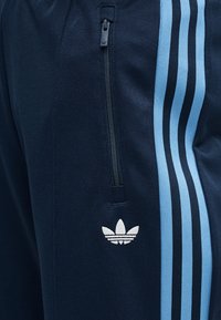 Μπλε σκούρο adidas φόρμα με ανοιχτό μπλε τριπλές ρίγες, κατακόρυφη τσέπη με φερμουάρ και λευκό λογότυπο trefoil κάτω από την τσέπη.