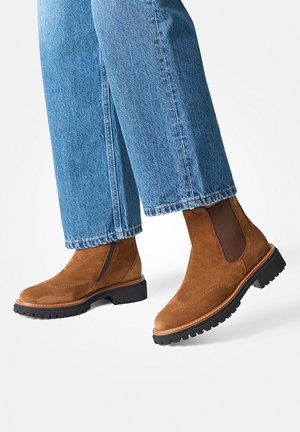 Bottines en suède marron avec semelles en caoutchouc, panneaux latéraux élastiques et surpiqûres décoratives, associées à un jean en denim bleu court.