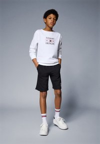 Fehér pulóver, rajta a "Tommy Hilfiger" logóval, fekete rövidnadrág, fehér sportcipő és csíkos zokni. Egyszerű dizájn, laza illeszkedés és a kényelemre összpontosít.