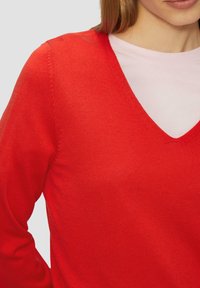 Czerwony sweter w serek o prążkowanej teksturze, dopasowane rękawy oraz nałożona różowa koszula, pokazująca czyste linie i wyraźny kontrast kolorów.