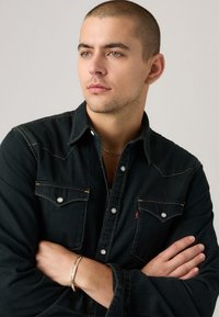 Dark denim shirt with a classic western design, featuring snap buttons, double chest pockets, and contrast stitching. Accessories include a bracelet.