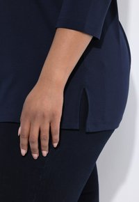 Marineblaue Tunika mit 3/4 Ärmeln und seitlichen Schlitzen, aus weichem Material. Die Hand, die auf der Hüfte ruht, zeigt eine glatte, natürliche Hautstruktur.
