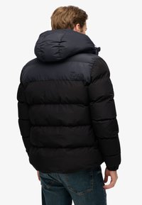 Czarna i ciemnoszara kurtka puchowa z kapturem, z pikowanym wzorem i logo "Superdry" na ramieniu.