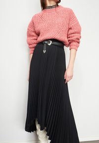 Rosa, strukturierte Strickjacke mit weitem Ausschnitt, kombiniert mit einem schwarzen, plissierten asymmetrischen Rock, der einen schwarzen Gürtel mit einer silbernen Schnalle hat.