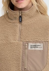 Gros plan sur une personne portant une veste en polaire beige avec deux zips et une poche poitrine portant l'inscription "DESIGNÉ AVEC DÉVOTION oxmo® EST. 2001."