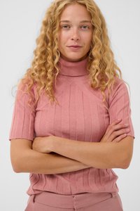 Kvinde med krøllet blonde hår iført en lyserød ribbet kortærmet turtleneck sweater og matchende bukser, med armene korslagte og en neutral ansigtsudtryk.