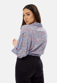 Light blue floral blouse with red and white patterns, collar, and puffed sleeves. Paired with black fitted pants highlighting the waist.