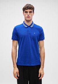 Királykék pólóing csíkos gallérral és rövid ujjal, bal mellen egy kis logóval és klasszikus szabással.