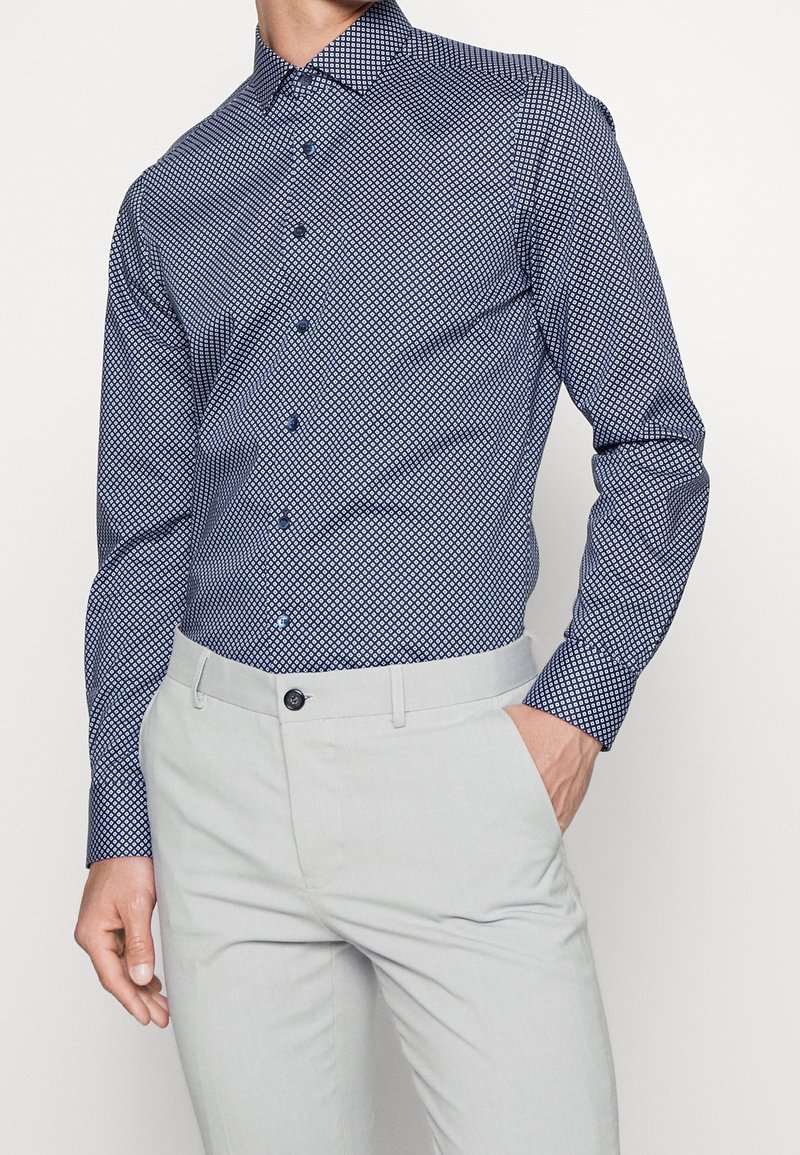 Camicia botton-down blu scuro con motivo geometrico; tessuto morbido e maniche lunghe. Abbinata a pantaloni grigi chiari su misura.