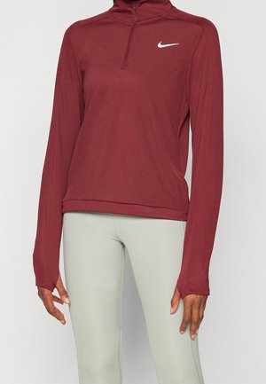 Osoba ubrana w bordową bluzę Nike z długim rękawem i suwakiem do ćwiartki oraz jasnoszare legginsy, stojąca na tle jednolitego tła.