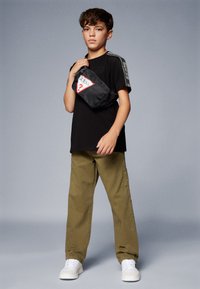 Svart t-skjorte med "Guess" logo, olivengrønne cargo-bukser og hvite joggesko. Modellen bærer en svart crossover-veske med rød og hvit logo.