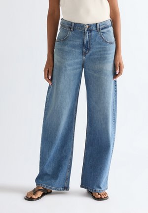 Γυναίκα που φοράει ανοιχτό μπλε τζιν παντελόνι με φαρδιά μπατζάκια και μαύρα σανδάλια με δέσιμο στο δάχτυλο, όρθια μπροστά σε απλό λευκό φόντο.