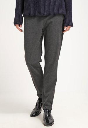 Person iført mørkegrå, tilspidsede bukser med elastiske manchetter, en navyblå ribstrikket sweater og skinnende sorte finere sko, stående mod en ensfarvet baggrund.