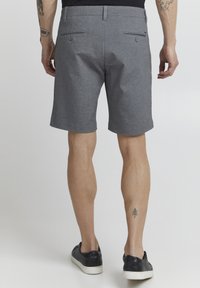 Shorts gris sur mesure en tissu texturé, dotés de deux poches arrière avec boutons et d'une coupe droite épurée, associés à des baskets noires.