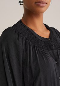 Közelkép egy személyről, aki fekete gombos blúzt visel, smockolt vállrésszel és nagy, kerek ezüst fülbevalókkal, semleges háttérrel.