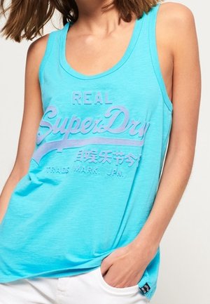 Frau trägt ein ärmelloses türkisfarbenes Tanktop mit dem Logo „Real SuperDry“ und japanischen Schriftzeichen, kombiniert mit weißen Hosen.
