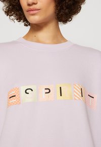 Femme portant un sweat-shirt lavande avec des blocs colorés à motifs formant le mot « ESPRIT » sur la poitrine.