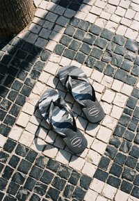 Ζευγάρι ασπρόμαυρα Havaianas σαγιονάρες με σχέδιο σε ασπρόμαυρο πλακόστρωτο δάπεδο με σκιά δέντρου.