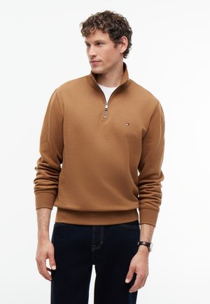 Hombre vestido con un pulóver marrón con cremallera hasta la mitad del pecho, con un pequeño logo en el pecho, pantalones oscuros y un reloj de pulsera, mirando hacia un lado.