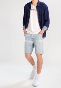 Shorts de mezclilla en azul claro con detalles desgastados, combinados con una camiseta blanca y una camisa de botones azul oscuro. Llevados con zapatillas blancas.