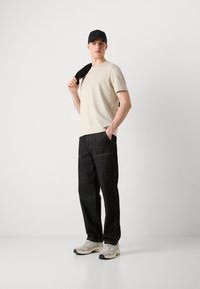 Άνδρας που φοράε ι μπεζ μπλουζάκι, μαύρο καπέλο, σκοτεινό παντελόνι, λευκά αθλητικά παπούτσια, κρατά μαύρο μπουφάν στον ώμο, στέκεται μπροστά σε απλό λευκό φόντο.