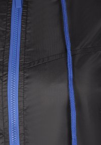 Giacca nera con zip blu e coulisse coordinata. Il tessuto è liscio con una leggera lucentezza e le linee di cucitura sono visibili.