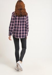 Námořnická a červená kostkovaná košile s dlouhými rukávy, spárovaná s černými skinny džíny a bílými teniskami, viděná zezadu.