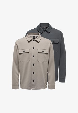 Due camicie a manica lunga con bottoni, colletto e tasche frontali con bottoni, una beige davanti a una grigio scuro, su sfondo bianco.